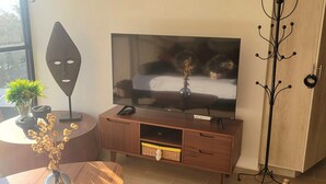 Un salon moderne avec un téléviseur posé sur un meuble en bois, une petite table avec un vase, un porte-manteau et une vue par la fenêtre.