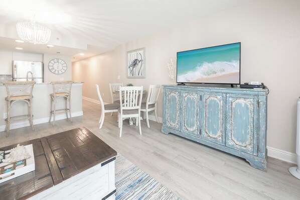 A modern kitchen with white cabinetry, a dining area with white chairs, a blue cabinet with artwork, and a large TV on the wall.