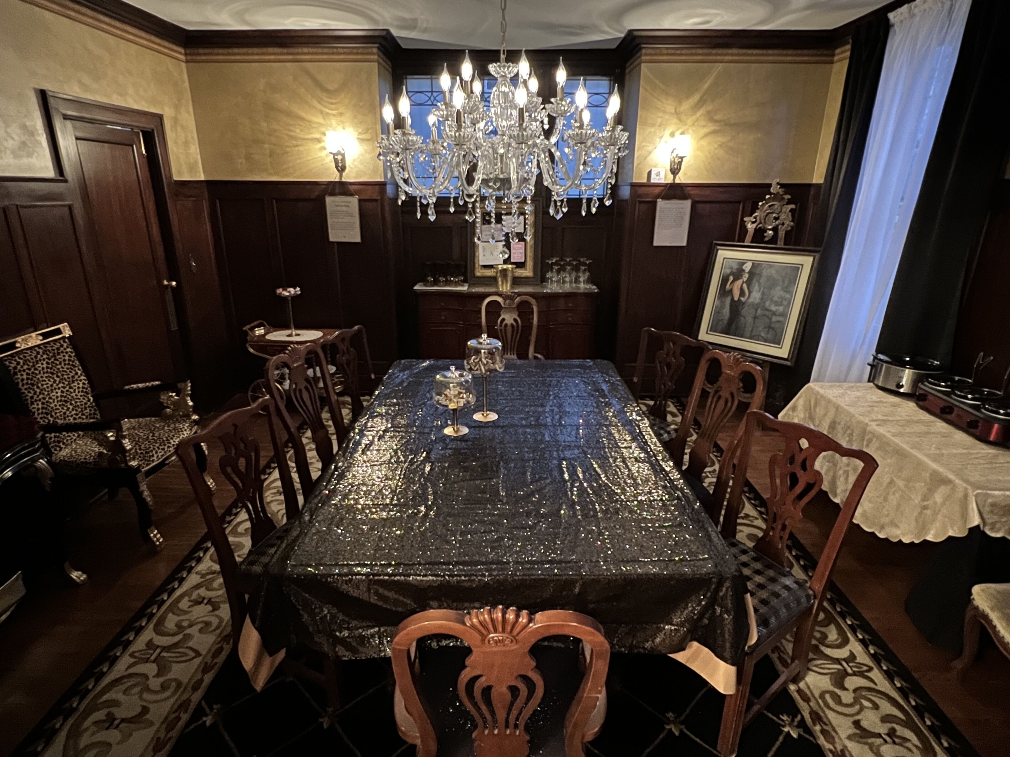 A formal dining room with a chandelier, wooden chairs, a table with a decorative tablecloth, and a framed picture on the wall.