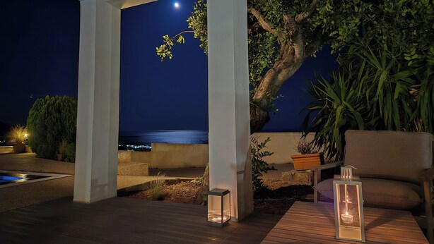 Ein Patio im Freien mit Blick auf das Meer bei Nacht, mit Holzbalkendecke, Topfpflanzen und zwei Laternen.