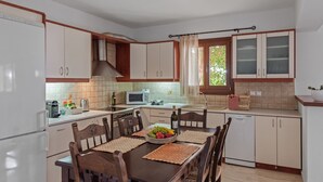 Eine Küche mit weißen Schränken, einem Holztisch mit Platzsets, einer Obstschale und einem Fenster mit Vorhängen.