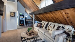 A loft-style room with a high ceiling, a couch with patterned cushions, a dartboard, and a gaming machine.
