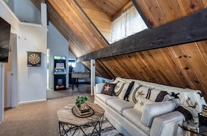 A loft-style room with a high ceiling, a couch with patterned cushions, a dartboard, and a gaming machine.
