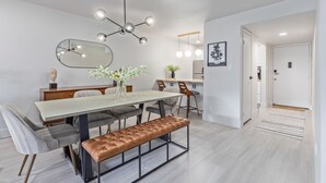 Een moderne eethoek met een lange tafel, gestoffeerde stoelen, een bank, een hanglamp, een spiegel en een lijstwerk met kunst.