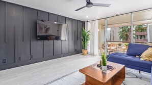 Een moderne woonkamer met een flatscreen televisie aan de muur, een plafondventilator, een blauwe bank en een houten salontafel met decoratieve objecten.