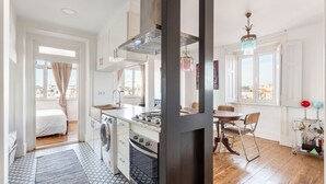 A compact kitchen with a stove, sink, and washing machine, a dining area with a table and chairs, and a view of the city through large windows. - JOIVY Cosy 2-bed flat w/desk in Arroios (Lisbon)