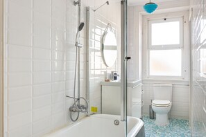 Ein Badezimmer mit weißen Fliesen, einer weißen Badewanne, einer Duschkabine aus Glas, einer Toilette und einem Fenster mit Ausblick.