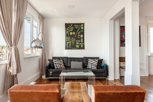 Ein modernes Wohnzimmer mit einem schwarzen Ledersofa, einem Glastisch und zwei braunen Sesseln. Ein botanisches Bild hängt an der Wand, und ein Fenster mit Vorhängen ist sichtbar.