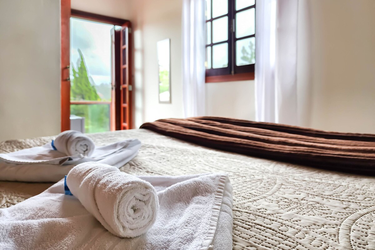 A bed with rolled towels, a window with curtains, and a view of greenery outside.