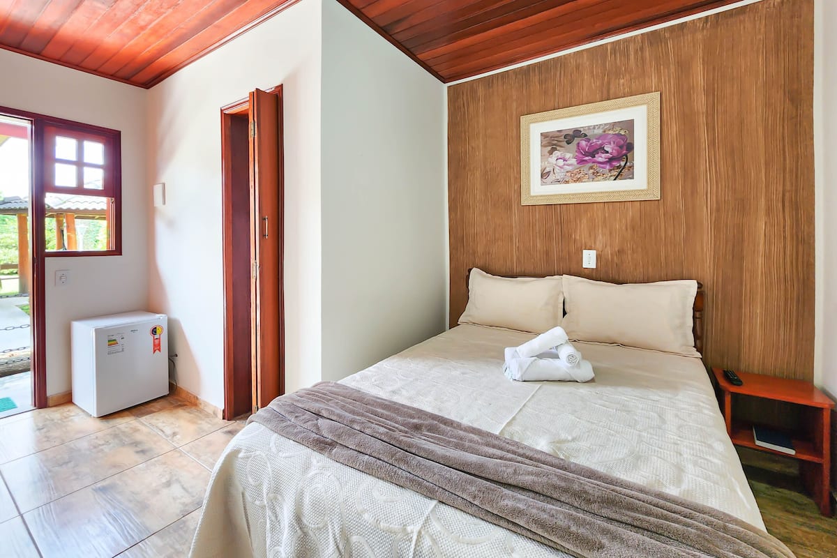 Ein Schlafzimmer mit Bett, einem kleinen Tisch, einem Bild an der Wand und einer Tür zu einem Außenbereich.