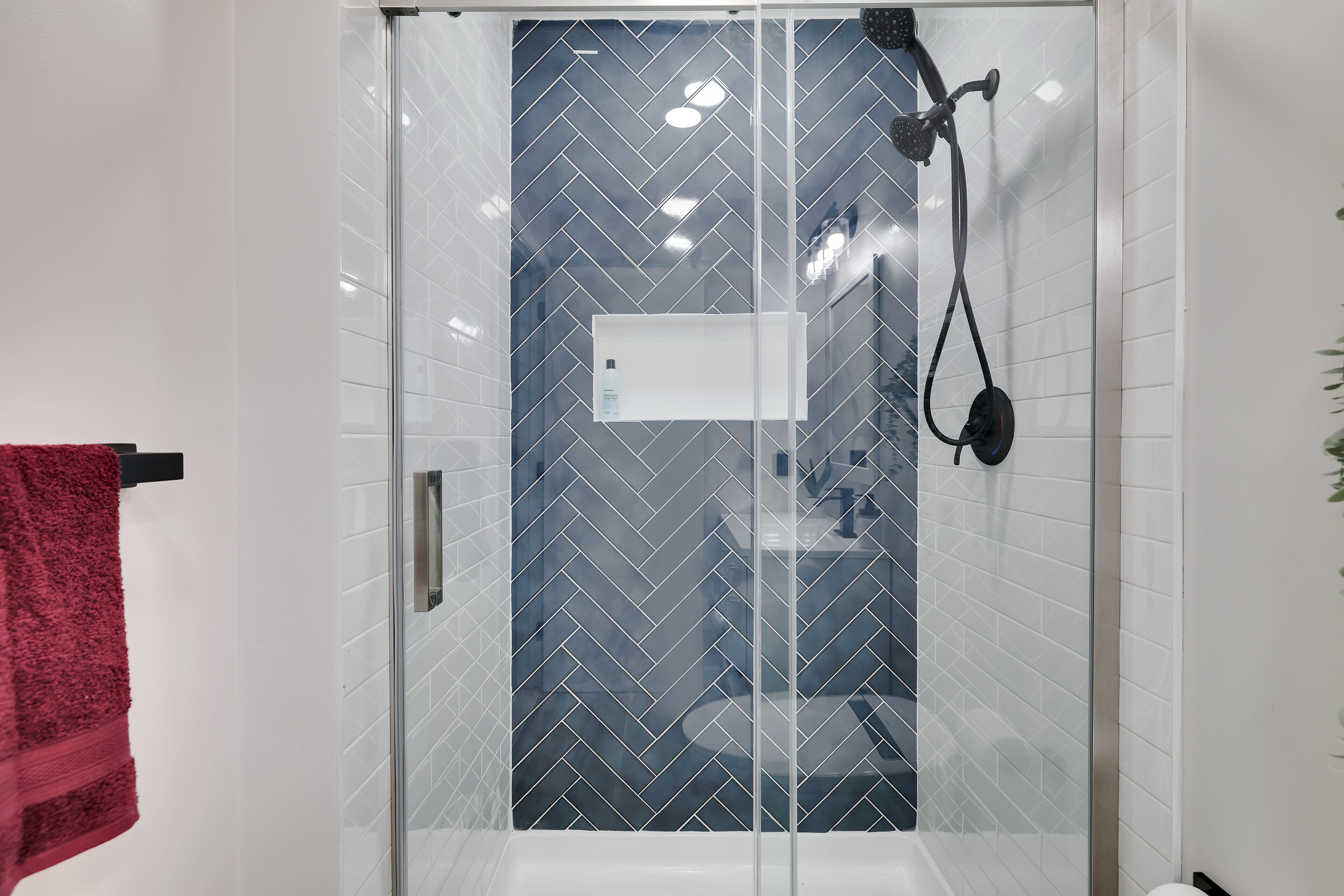 A glass-enclosed shower with a herringbone tile pattern, a visible bottle of shampoo, and a red towel on a rack.
