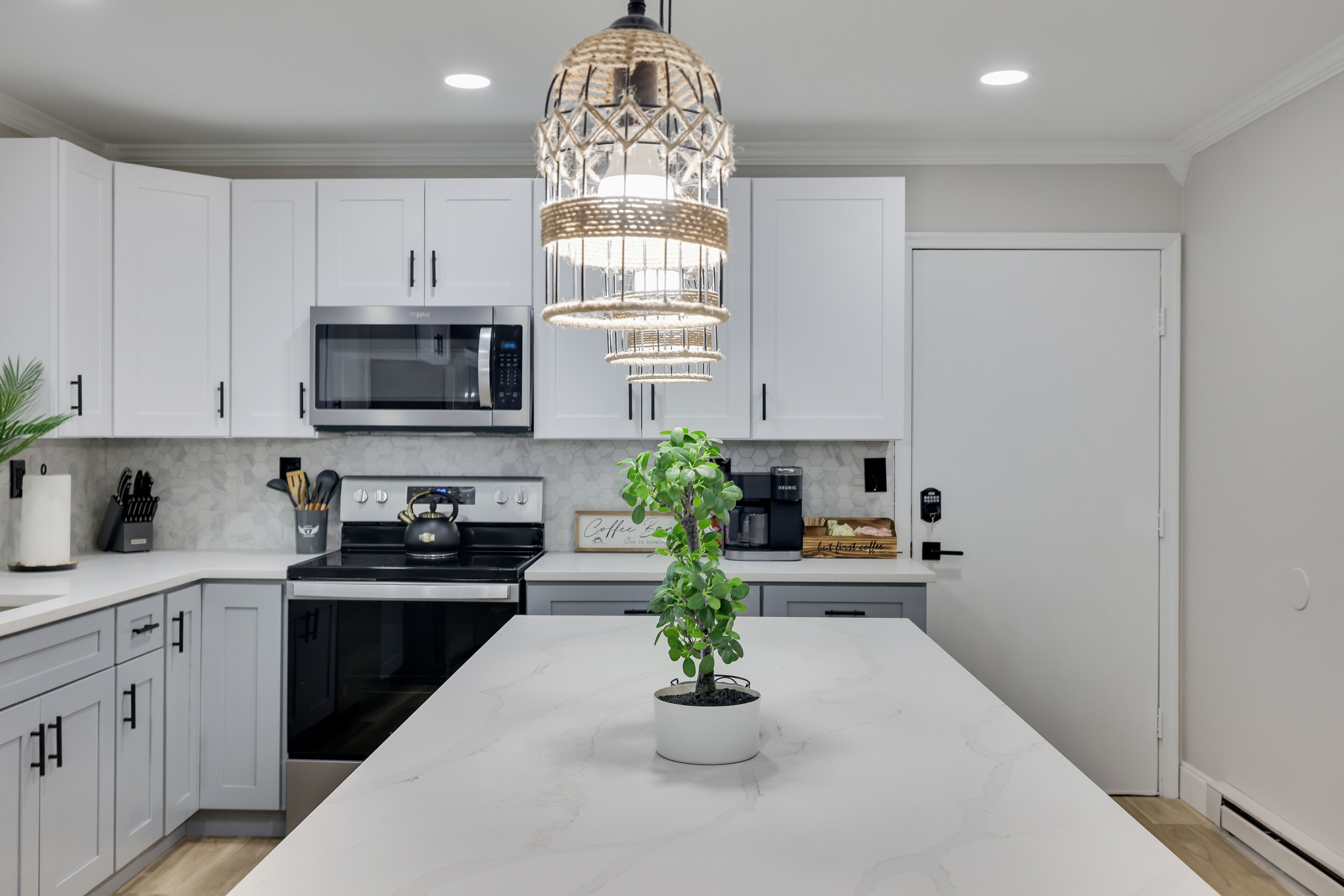 A modern kitchen with white cabinets, a central island with a potted plant, a stainless steel microwave, and a hanging light fixture.