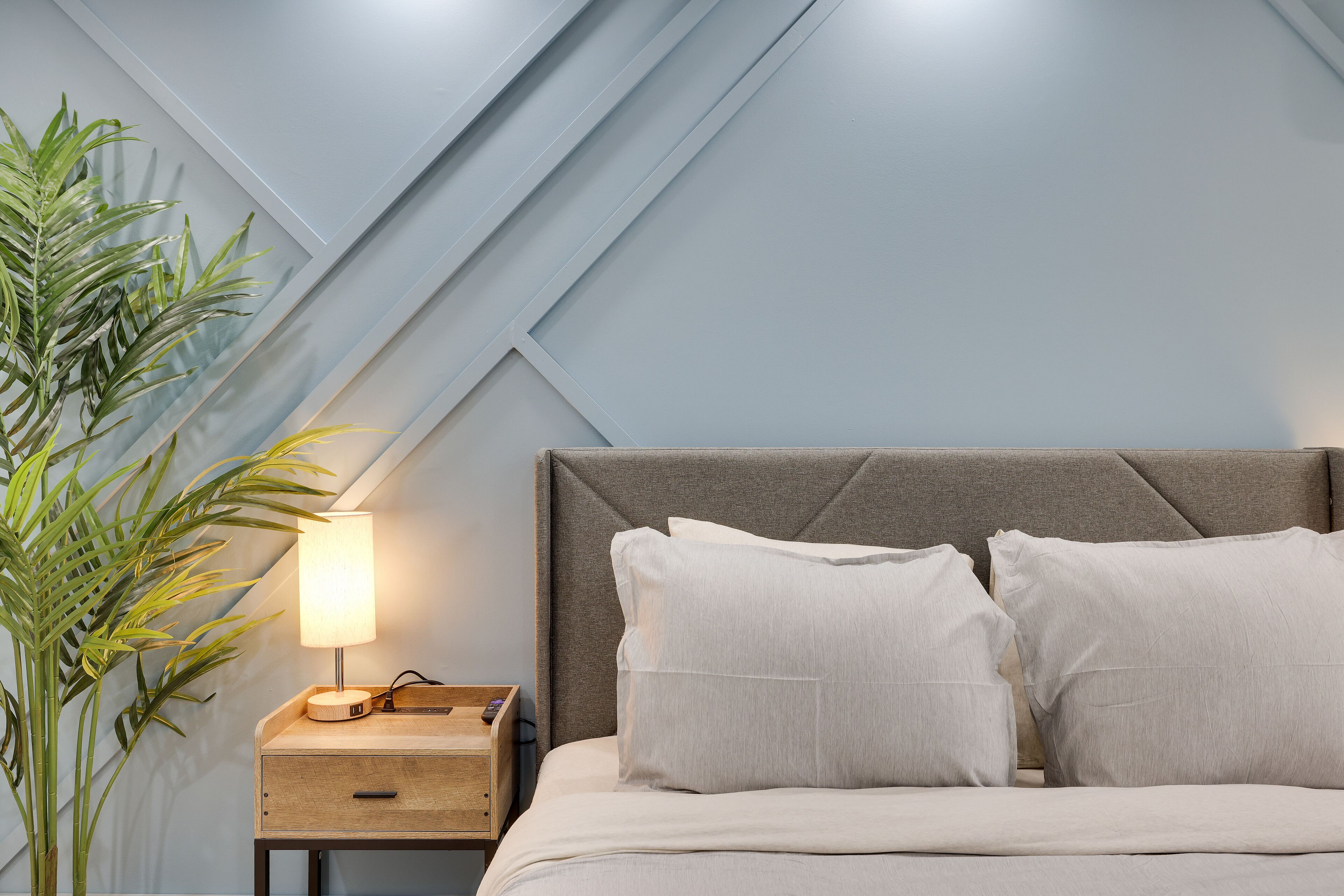 Una cama bien hecha con cabecera gris, una mesita de noche con lámpara y una planta en maceta.