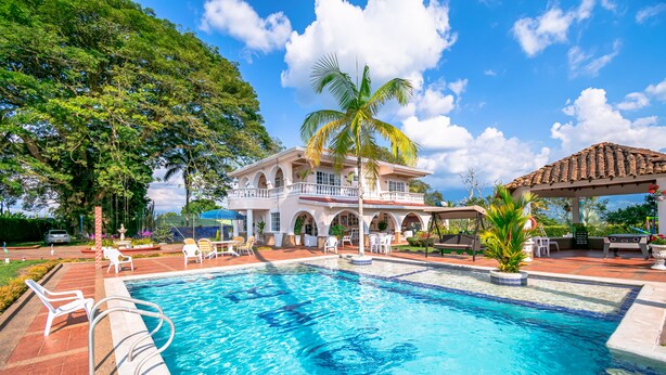 Una piscina con sillas de descanso y un pabellón, rodeada por una casa grande con balcón y palmeras.