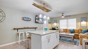 Een moderne keuken met een centraal eiland, witte kasten en hanglampen. Er is een zithoek met een houten bank en een sofa met blauwe kussens.