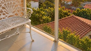 A balcony with a white wrought-iron bench, overlooking a residential area with red-tiled roofs and a sunset view.