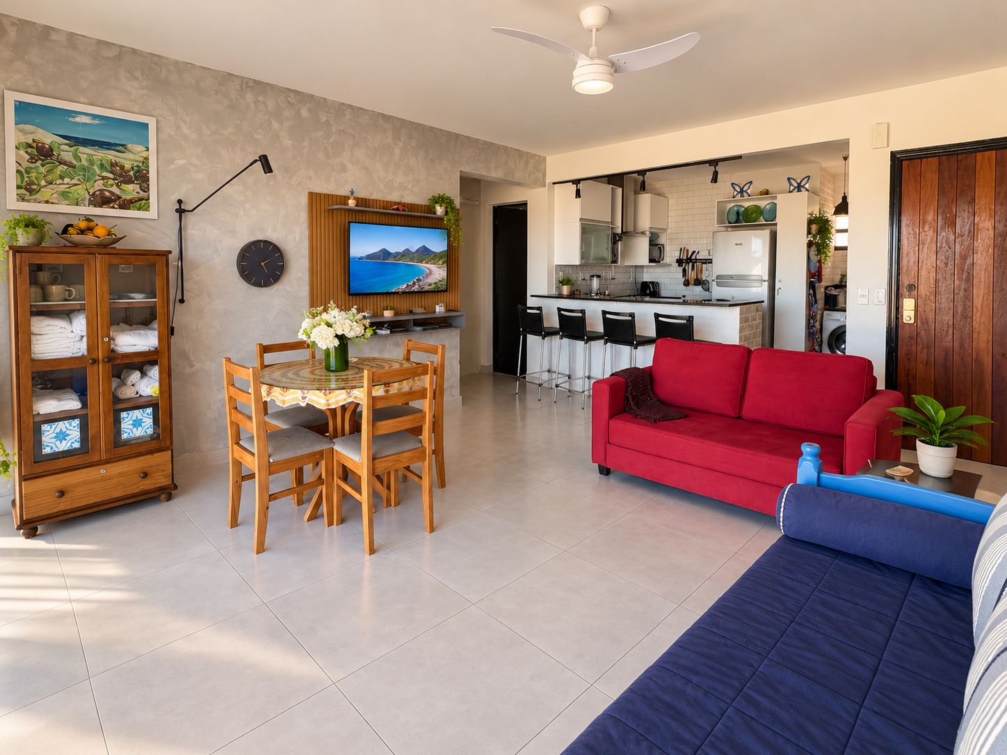Un soggiorno con un divano rosso, una poltrona blu, una zona pranzo con tavolo e sedie, una cucina sullo sfondo e una ventola a soffitto.