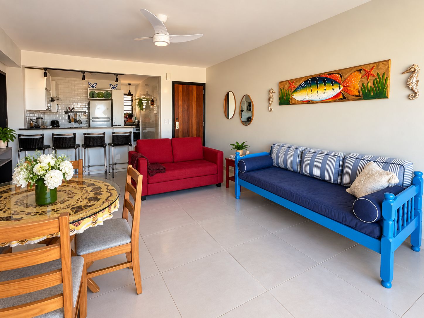 A living room with a red sofa, a blue sofa, a dining table with chairs, a kitchen in the background, and a ceiling fan.