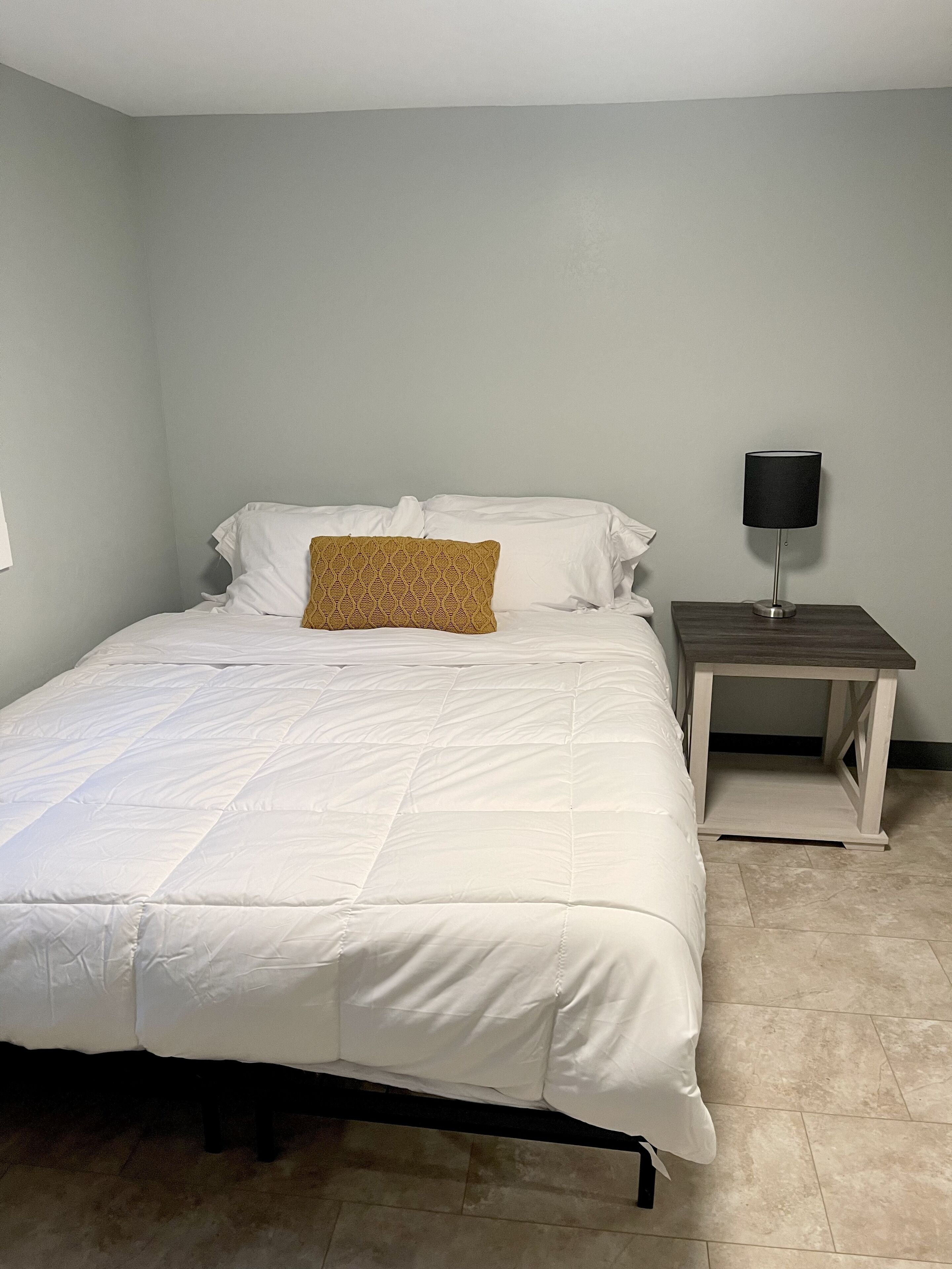 Un lit bien fait, avec une literie blanche et un seul coussin à motifs, une table de chevet avec une lampe et un mur sobre.