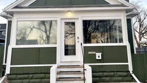 A small, single-story house with a green exterior and white trim, featuring a front door with a glass panel and a small porch with steps. - NEW! Heart of Downtown: The Hill Oasis (Grand Junction)
