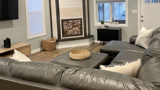A modern living room with a grey leather sofa, a fireplace with a stone surround, a flat-screen TV mounted on the wall, and a window with a 'Welcome' sign. - NEW! Heart of Downtown: The Hill Oasis (Grand Junction)