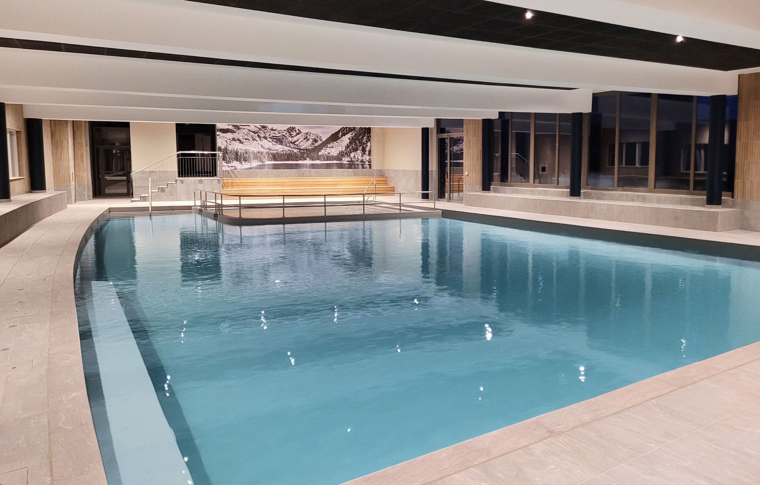 A large indoor swimming pool with clear blue water, a wooden bench, and a mountain view mural.