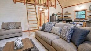 Ein gemütliches Wohnzimmer mit einer Holztreppe, einer Couch mit gemusterten Kissen und einem Küchenbereich im Hintergrund.