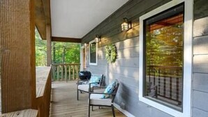Eine überdachte Veranda mit Holzboden, zwei Stühlen, einem Grill und großen Fenstern mit Blick auf Grünflächen.