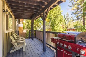 A covered deck with Adirondack chairs, a barbecue grill, and a view of trees.