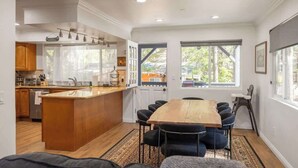 Une cuisine moderne dotée d’un îlot central, d’un coin repas avec une grande table et des chaises en bois, et de grandes fenêtres offrant une vue sur l’extérieur.