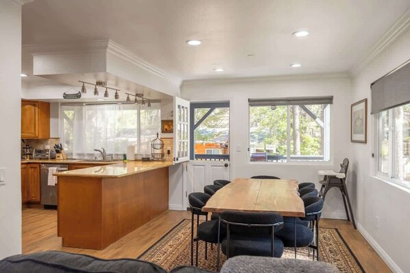 A modern kitchen with a central island, a dining area with a large wooden table and chairs, and large windows with a view of the outdoors.