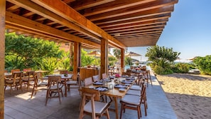 An outdoor dining area with wooden tables and chairs, a covered roof, and a view of the beach and ocean.