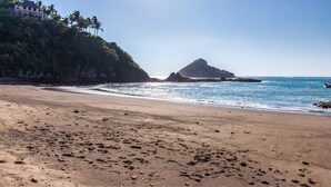 Ein Strand mit Fußabdrücken, die zum Wasser führen, ein Felsen und ein Gebäude mit einem Balkon an einem Hügel.