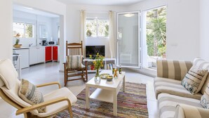 Een moderne woonkamer met een bank, fauteuil en salontafel, met een keuken op de achtergrond en een schuifpui die uitkomt op een balkon.