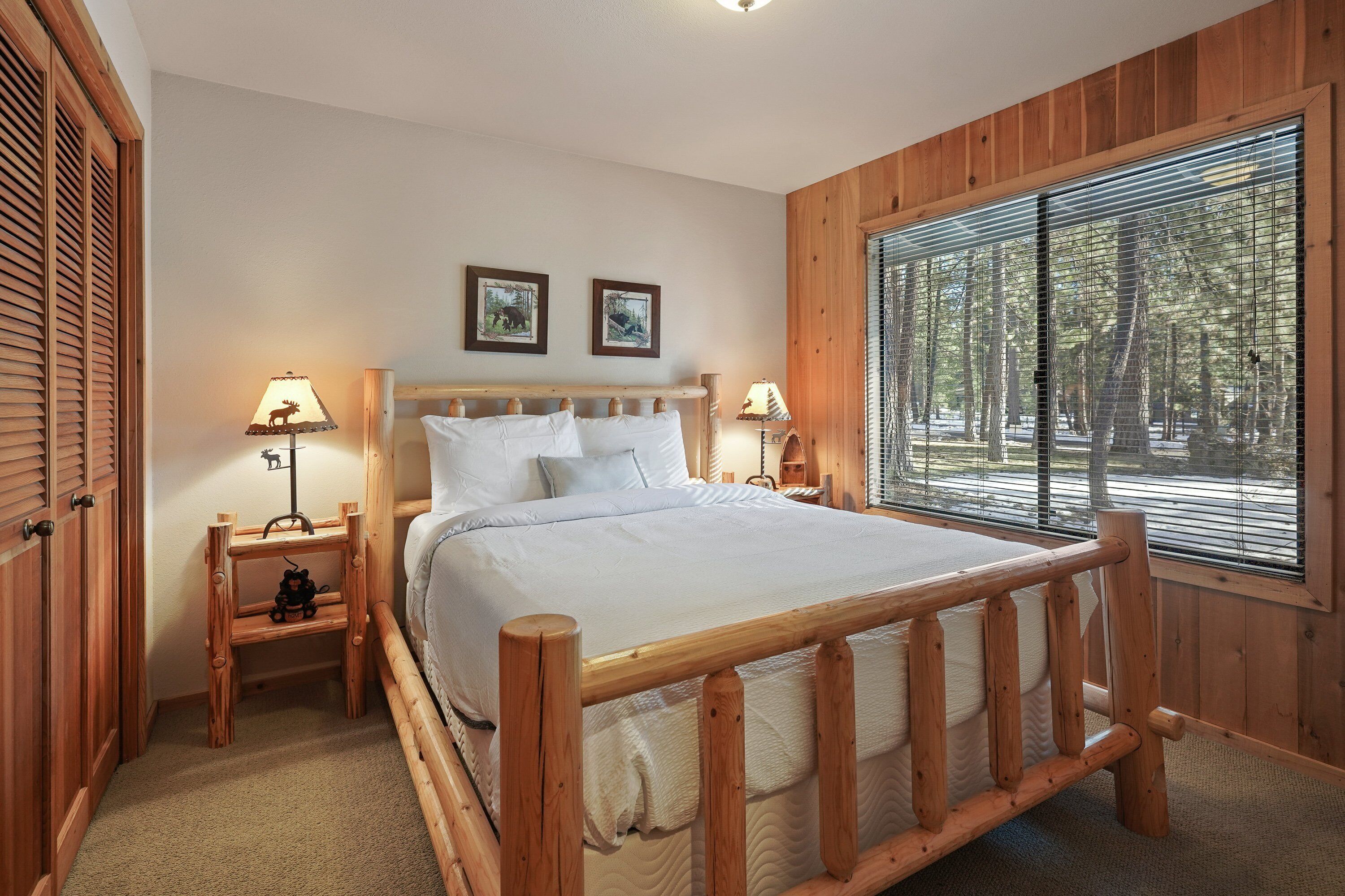 A wooden-framed bed with white bedding, two bedside tables with lamps, and a window with blinds showing a forest view.
