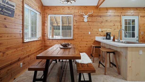 Interior de uma cabana de madeira com uma mesa, bancos, área de cozinha e um cabeça de veado montada na parede.