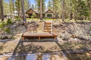 A wooden deck with chairs and a small table, surrounded by a forested area with a house in the background. - 110 Crane - Spectacular River Front + Access, Hot Tub, Ping Pong, South of Sunriver Resort (Sunriver)