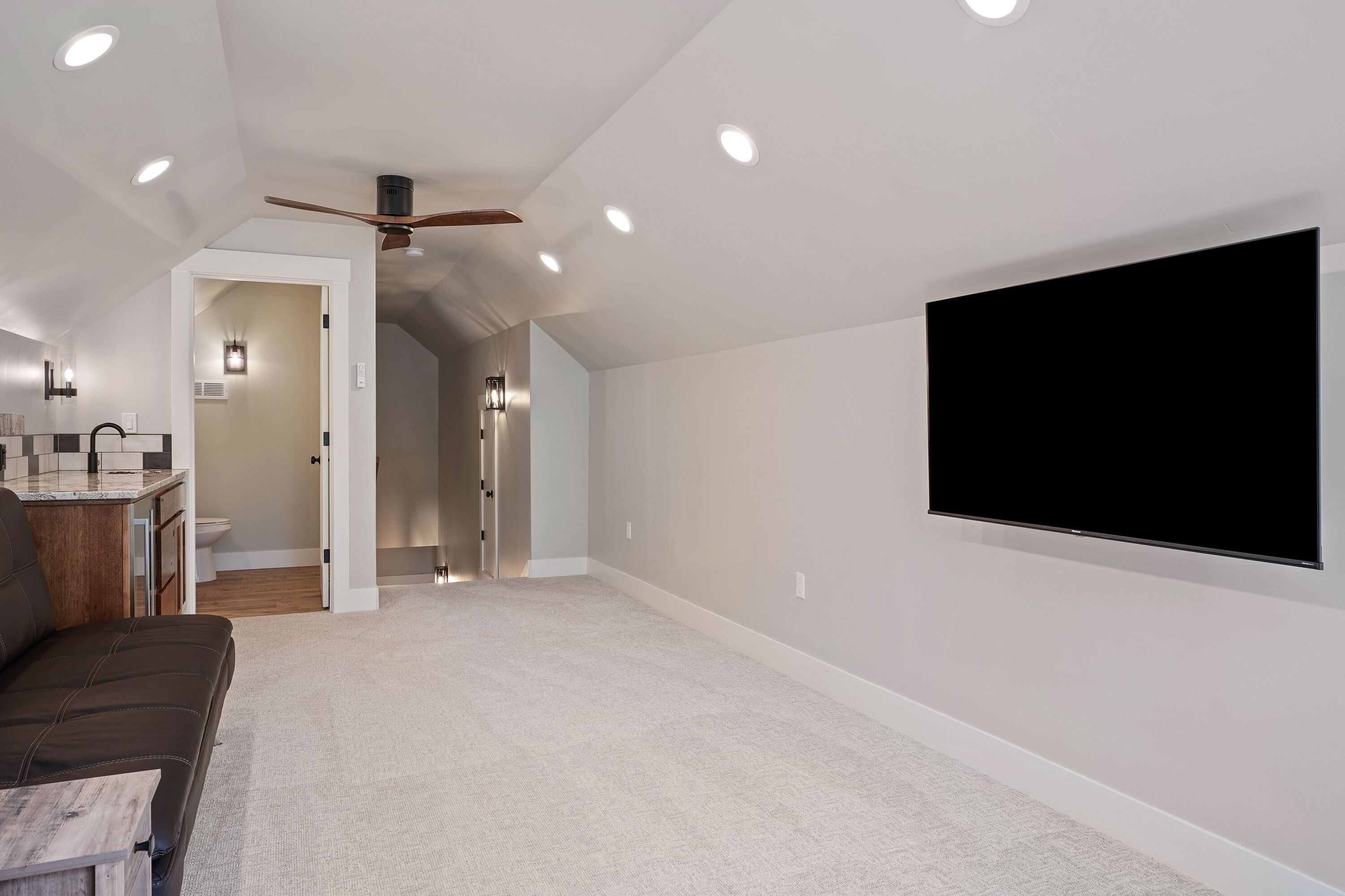 A spacious room with a ceiling fan, recessed lighting, a flat-screen TV mounted on the wall, and a carpeted floor.