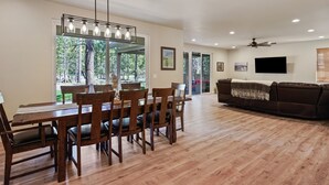 A spacious living area with a wooden dining table and chairs, a brown leather sofa, and a flat-screen TV mounted on the wall. - 110 Crane - Spectacular River Front + Access, Hot Tub, Ping Pong, South of Sunriver Resort (Sunriver)