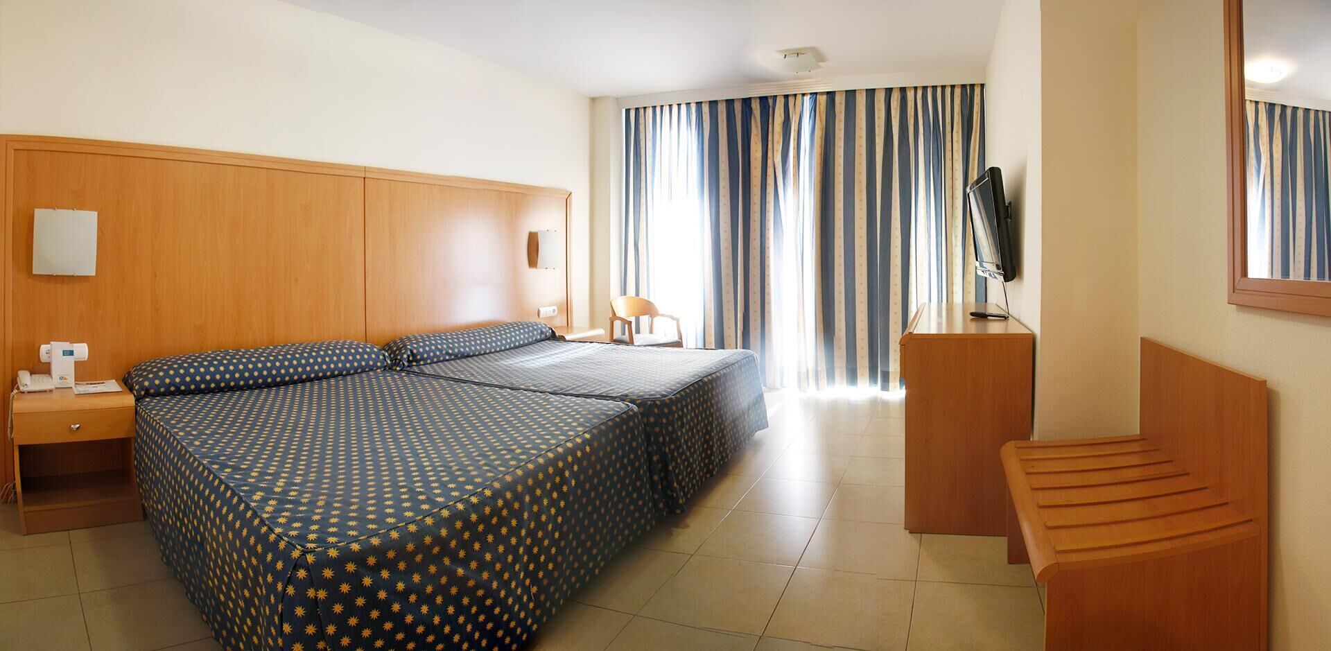 A hotel room with a large bed, a desk, a chair, a TV, and a window with curtains.