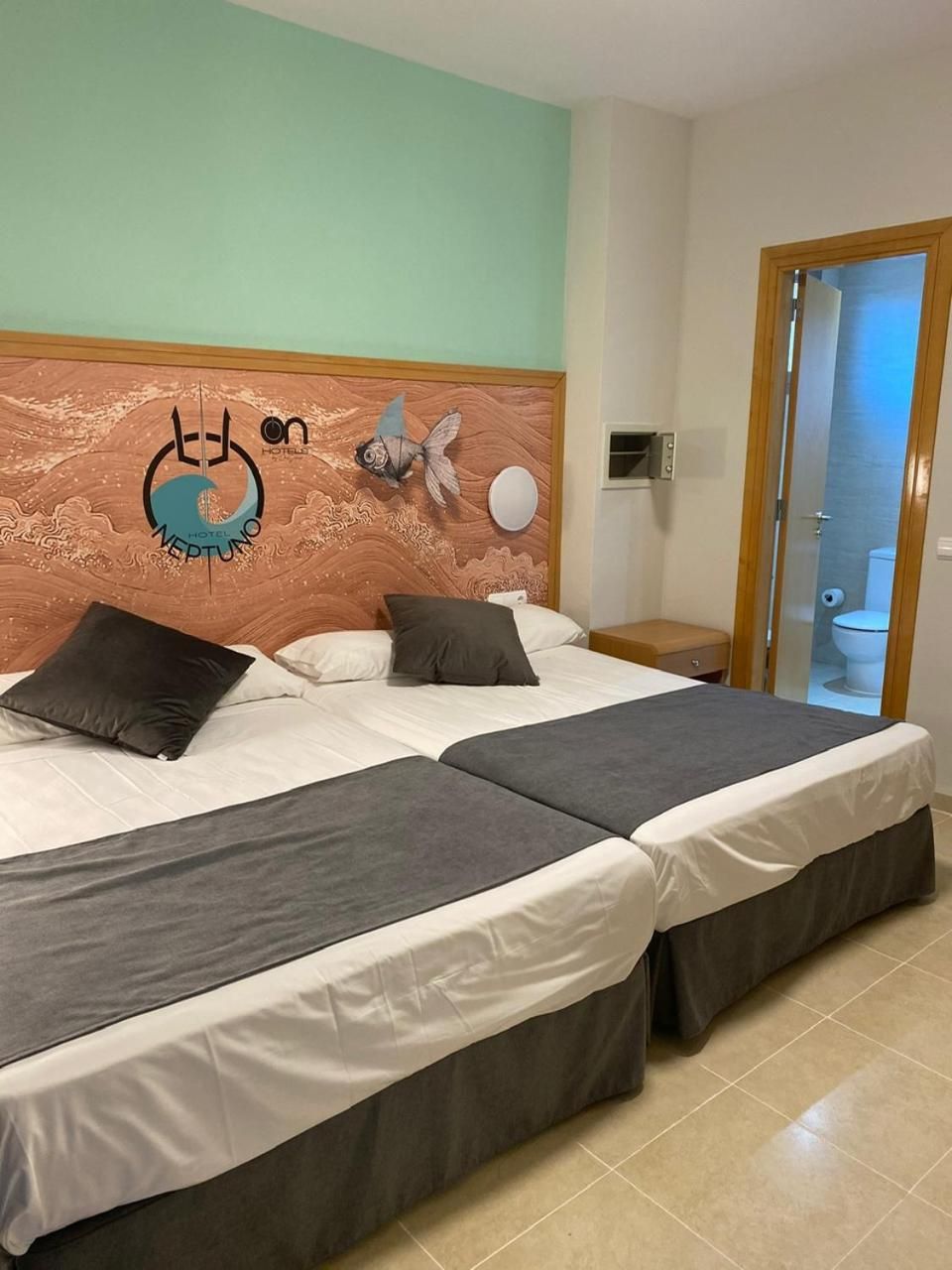 Una habitación de hotel con dos camas, cabecera de madera con diseño de mariposa y luna, una mesita de noche pequeña y vista al baño a través de una puerta de cristal.