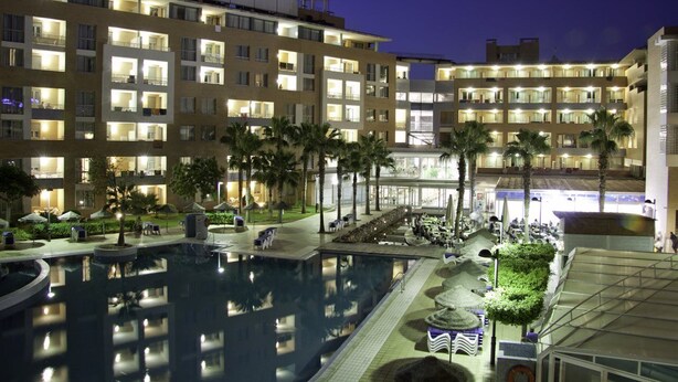Un hôtel à étages, doté d’une piscine extérieure, de palmiers et de balcons bien éclairés.