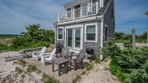 Una casa de playa de dos pisos con balcón, mobiliario exterior y una parrilla.