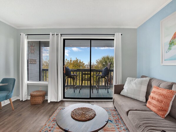A living room with a sofa, a coffee table, a wicker basket, and a view of the outdoors through a sliding glass door.