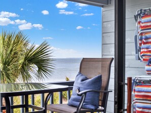 A balcony with a view of the ocean, a chair, a palm plant, and beach towels hanging on the wall.