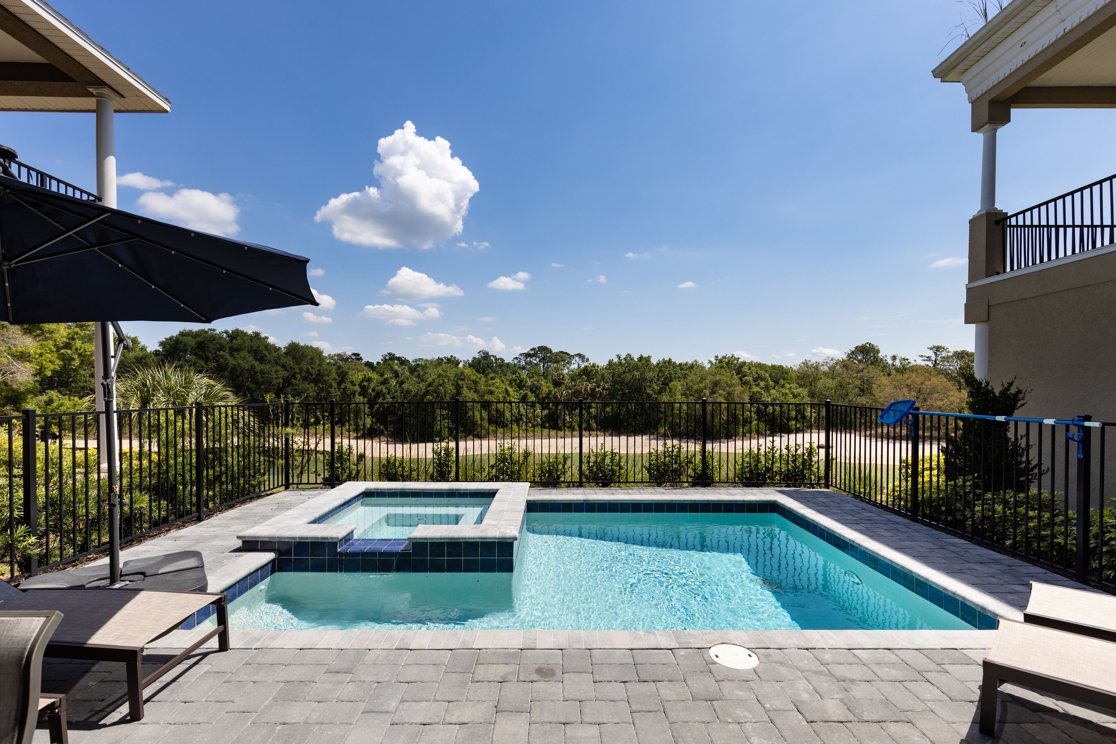 A rectangular swimming pool with a small hot tub, surrounded by a black metal fence, and a covered patio area with a table and chairs.