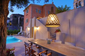 Un espace aménagé au bord de la piscine, avec une table et des chaises en bois, un luminaire suspendu et un escalier menant à un niveau supérieur.