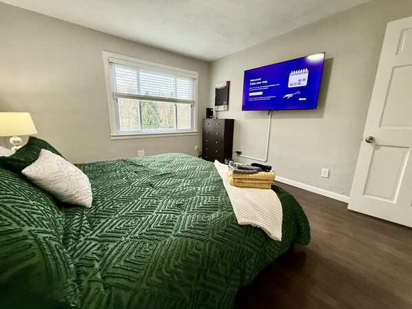 A bedroom with a bed, a TV mounted on the wall, a window with blinds, and a door.