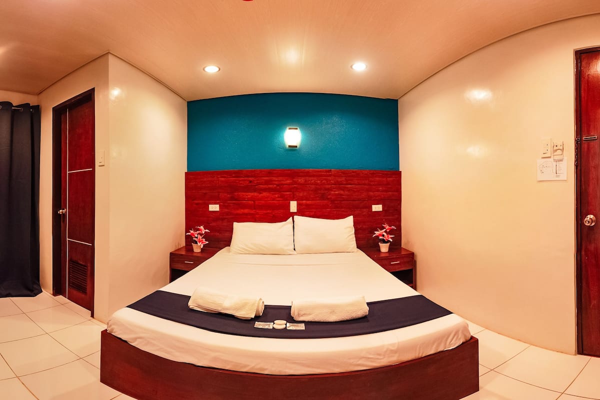 A hotel room with a curved bed, a red accent wall, a nightstand with a lamp, and a door with a red handle.