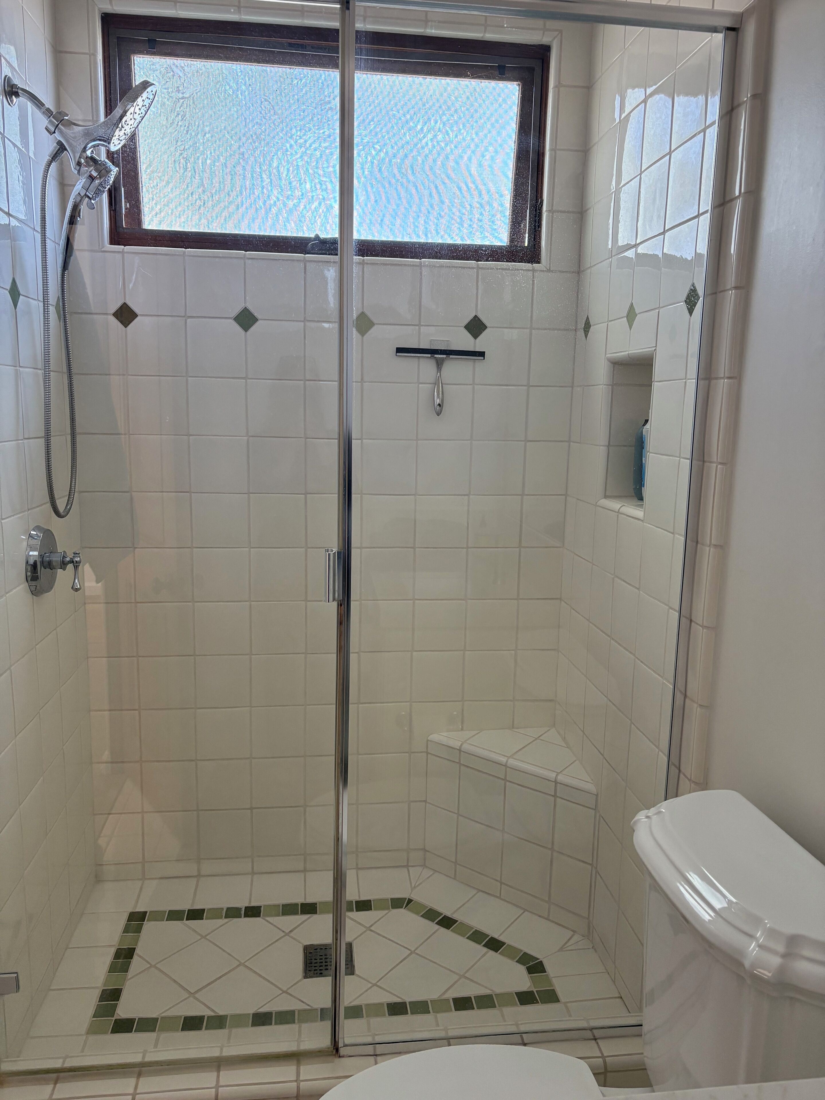 Um chuveiro com porta de vidro, uma janela acima e um vaso sanitário com tampa branca.